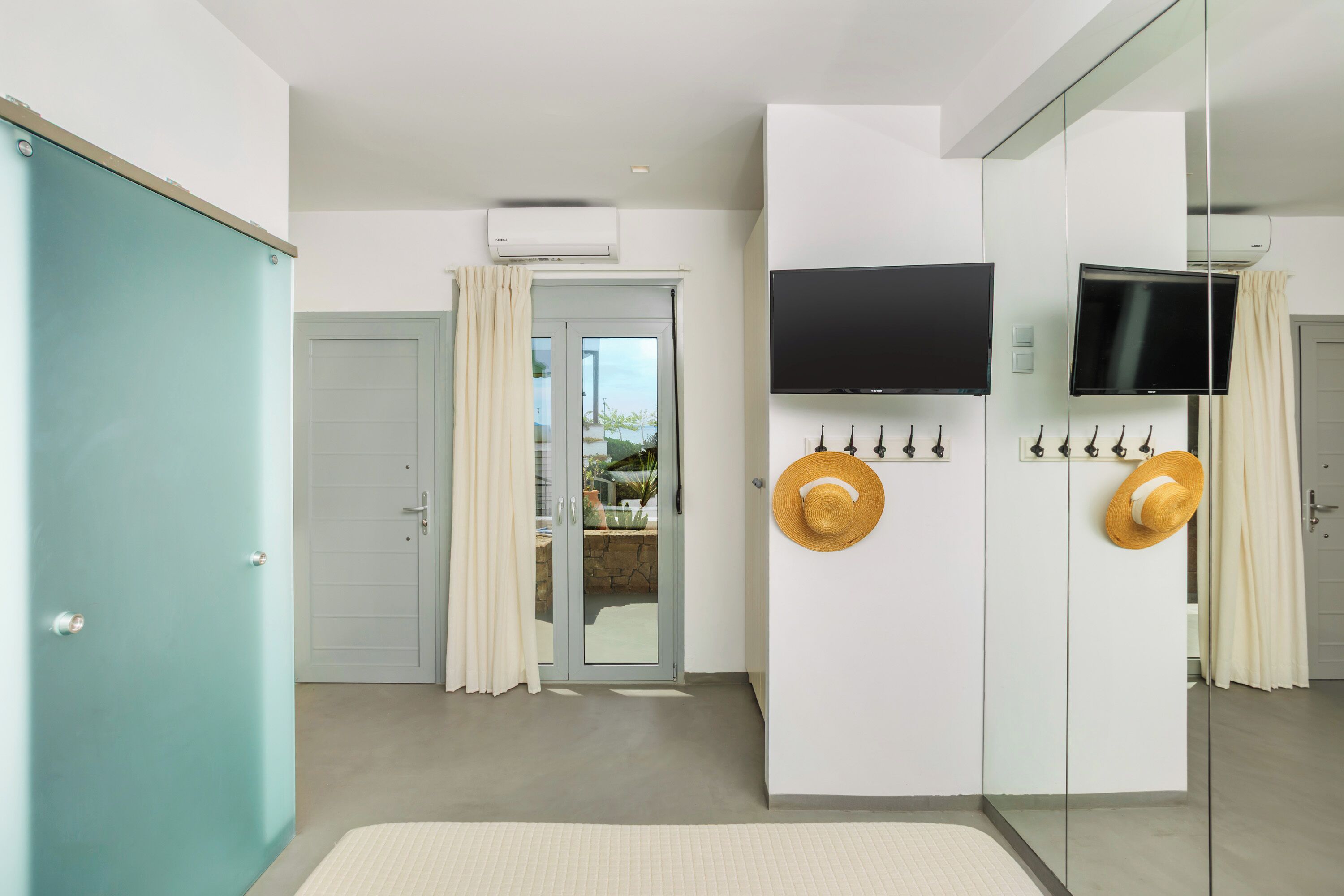A modern hotel room with a sliding glass door leading to a balcony, a flat-screen TV mounted on the wall, and a wardrobe with a mirror.
