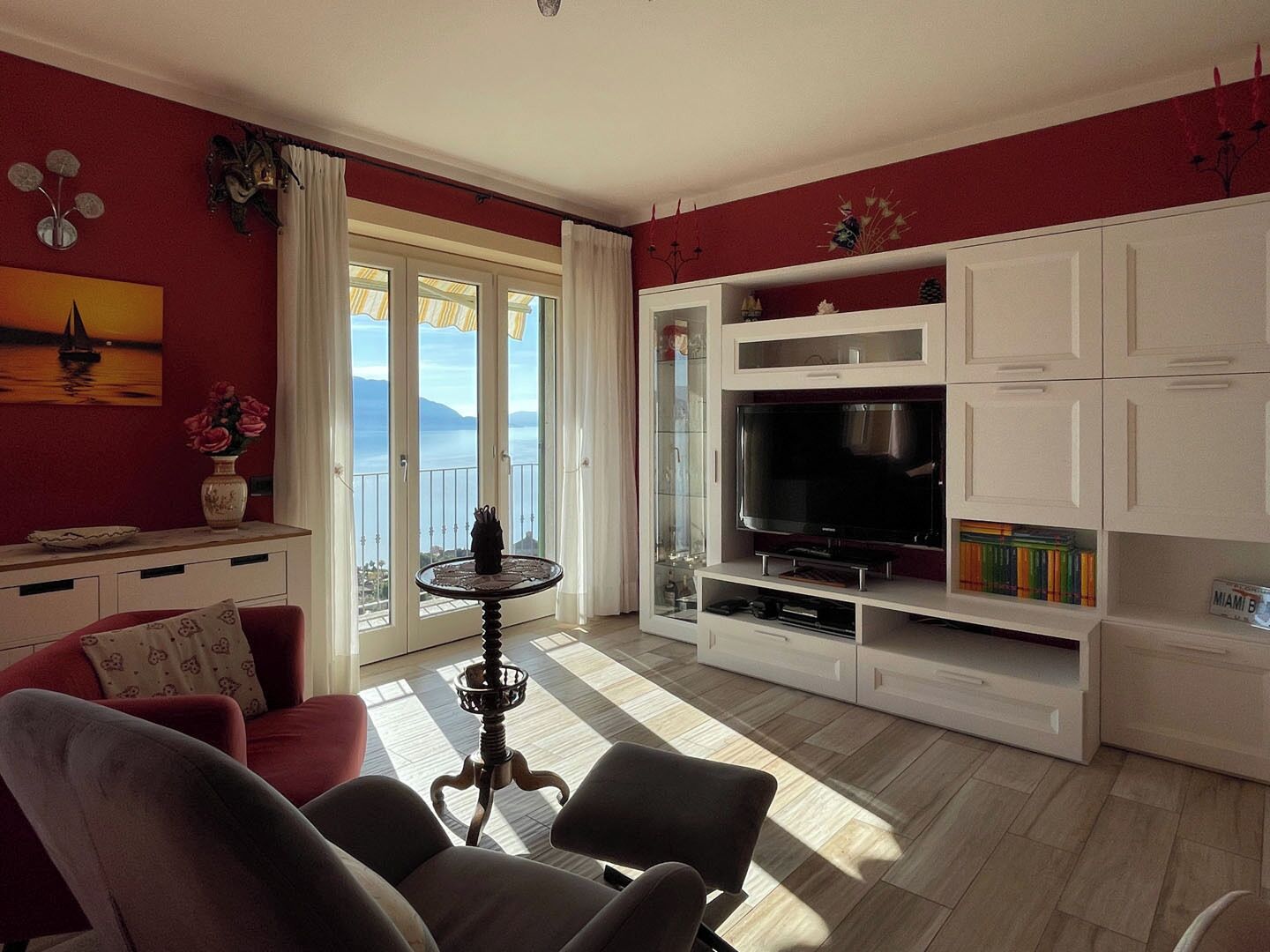Een woonkamer met een televisie, witte kasten, een rode wand en een balkon met gordijnen.