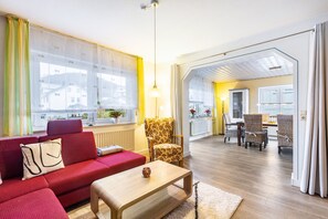 Een moderne woonkamer met een rode bank, een houten salontafel en een fauteuil met een patroon. Er is een eethoek met rieten stoelen en een tafel.