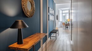 A hallway with a wooden console table, a black lamp, and a decorative mirror. There is a chair and a table with a plant in the background. - Ando Living - Abrantes Flats - 13 (Lisbon)