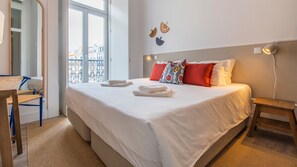 A neatly made bed with white linens, a small bedside table, and a chair with a blue frame. - Ando Living - Abrantes Flats - 23 (Lisbon)