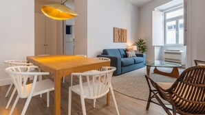 A modern living room with a wooden dining table, white chairs, a blue sofa, and a wicker chair. - Ando Living - Abrantes Flats - 23 (Lisbon)