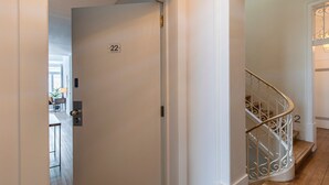 Un interior moderno con una puerta blanca identificada con el número '22', una escalera con barandillas de metal ornamentadas y un vistazo a un comedor con luz natural.