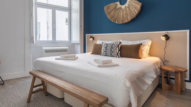 Una cama bien hecha con almohadas y un banco, una lámpara de pared y una ventana con persianas.