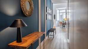 Un pasillo con una consola de madera, una lámpara negra, un espejo decorativo y vista hacia un área de comedor con una mesa y sillas.