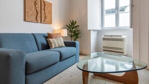 A living room with a blue sofa, a glass coffee table, a lamp, a plant, and a window with an air conditioner. - Ando Living - Abrantes Flats - 13 (Lisbon)