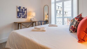 A hotel room with a large bed, a desk, a chair, a mirror, and a view of buildings through a window. - Ando Living - Abrantes Flats - 13 (Lisbon)