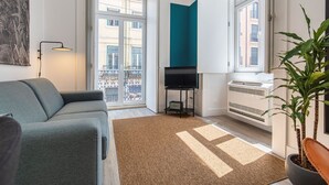 Un salon moderne avec un canapé gris, un téléviseur à écran plat posé sur un meuble, une grande fenêtre donnant sur des immeubles, un radiateur et une plante en pot.