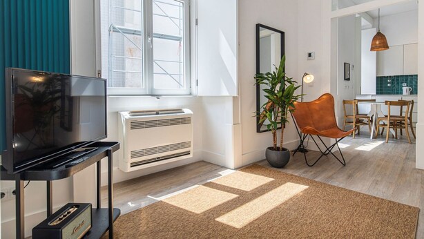 A modern living room with a flat-screen TV, a white radiator, a potted plant, a brown leather chair, and a dining area with wooden chairs and a table. - Ando Living - Abrantes Flats - 31 (Lisbon)