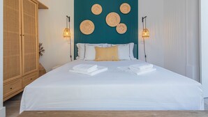 Un lit bien fait, avec du linge de lit blanc et deux serviettes pliées, un mur d’accent de couleur turquoise orné de motifs circulaires tissés, et une armoire en bois.