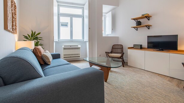A modern living room with a grey sofa, a glass coffee table, a wicker chair, a TV on a wooden stand, and a small shelf with books. - Ando Living - Abrantes Flats - 33 (Lisbon)