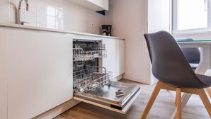 A kitchen with a dishwasher, a coffee maker, and a chair. - Ando Living - Abrantes Flats - 33 (Lisbon)