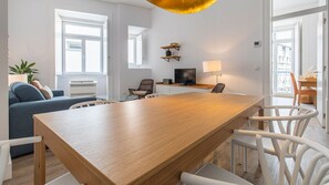 A modern living room with a wooden dining table, white chairs, a blue sofa, and a TV on a shelf. - Ando Living - Abrantes Flats - 33 (Lisbon)