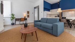 A modern living room with a blue sofa, a wooden coffee table, and a desk with a chair. There is a kitchen area with a dining table and chairs visible through an open doorway. - Ando Living - Abrantes Flats - 32 (Lisbon)