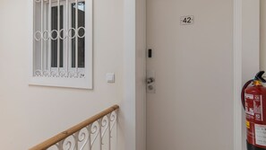 Uma porta com placa de identificação, um extintor de incêndio, uma janela com grades decorativas e um interruptor de luz.