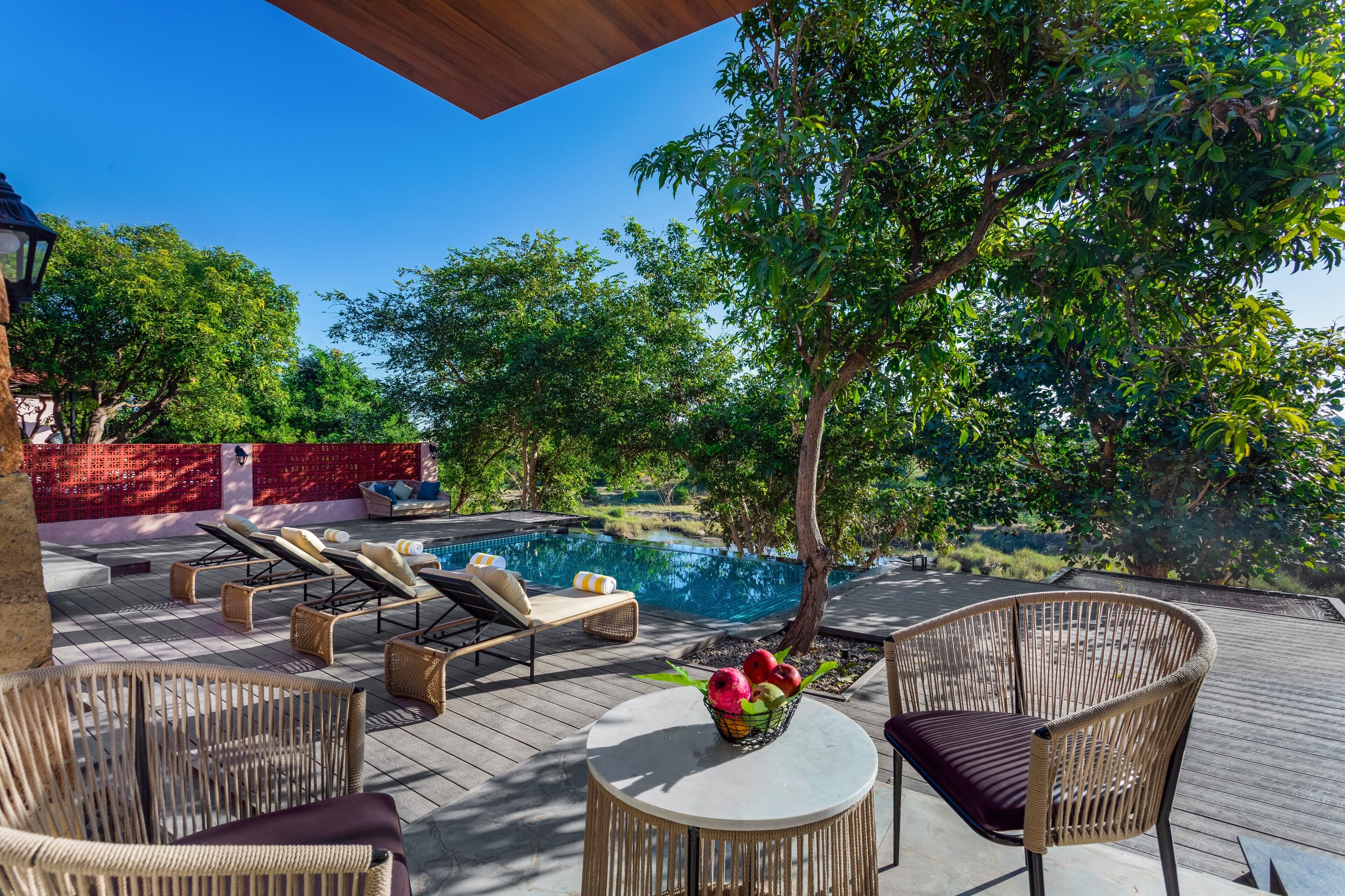 A poolside area with wicker furniture, a round table with a vase of flowers, and a view of a swimming pool surrounded by trees.