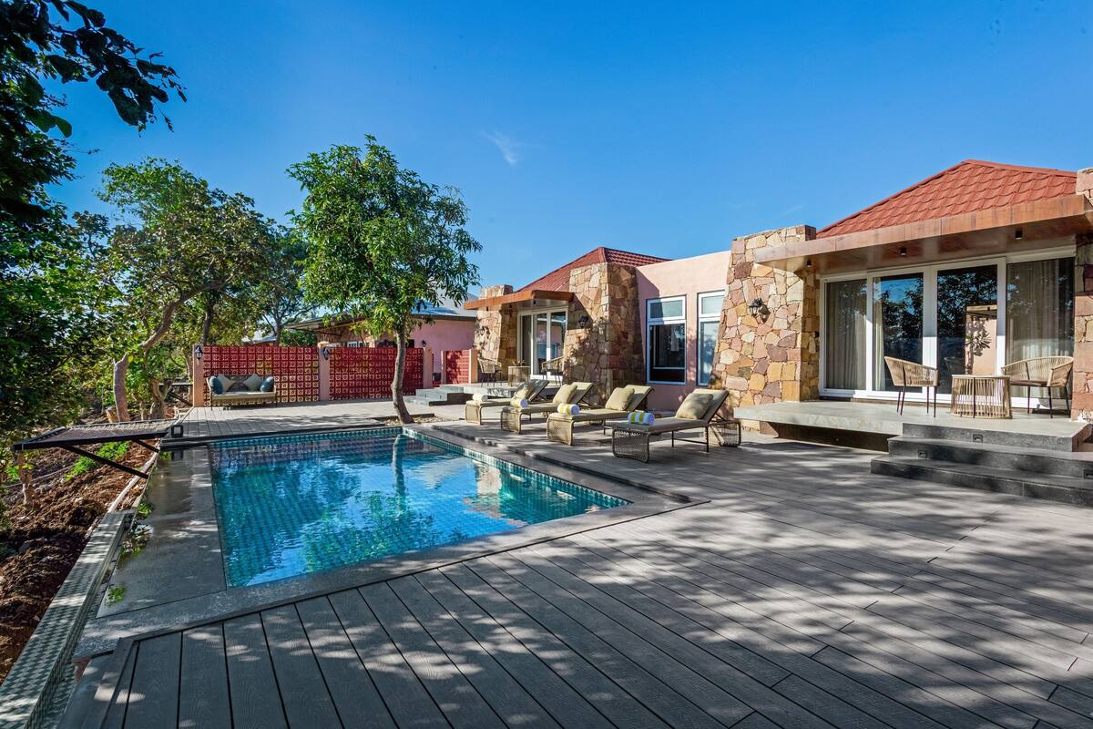 A pool area with a wooden deck, lounge chairs, and a stone building with a red roof.