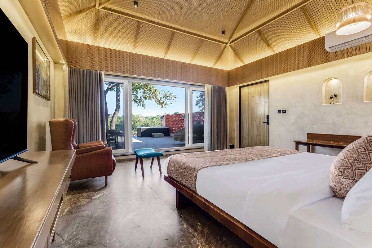 A hotel room with a large bed, a desk, a chair, a small table, and a sliding glass door leading to an outdoor area.