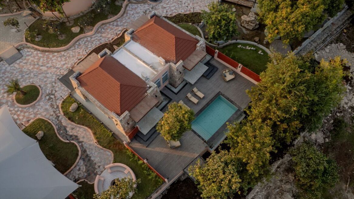 An aerial view of a property with a swimming pool, two buildings with red roofs, and landscaped gardens.