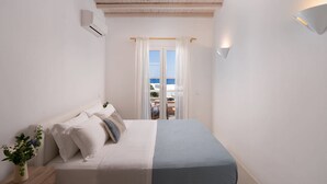 Ein Schlafzimmer mit Bett, Nachttisch, Topfpflanze und Meerblick durch ein Fenster.