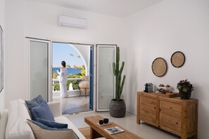 A modern living room with a sofa, coffee table, and a wooden cabinet. A large cactus plant is on the floor, and there are two woven wall decorations. - Villa Ioan with pool - My Rental Homes (Mykonos)