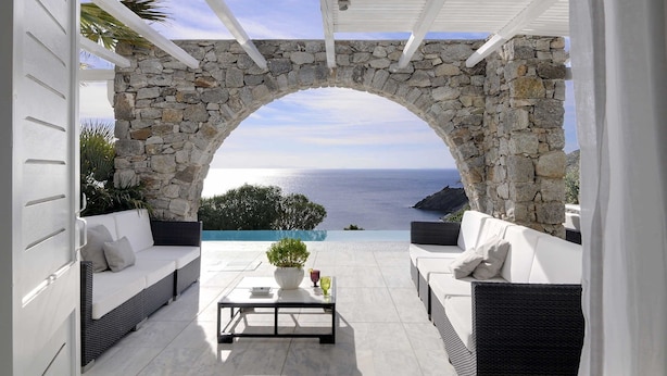 Ein steiner Torbogen führt zu einer Terrasse mit weißer Möbel und Blick auf das Meer.