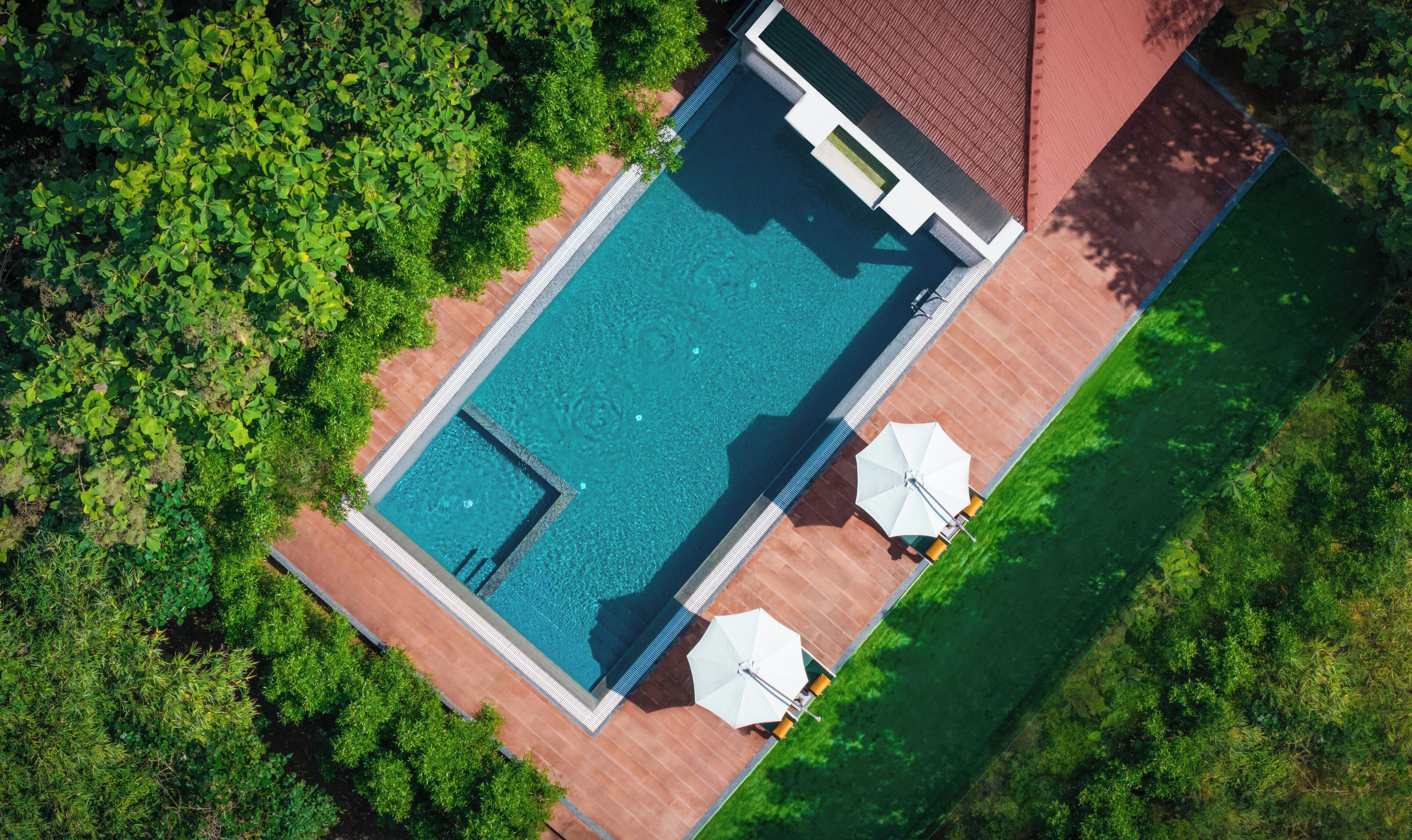 Vue aérienne d’une piscine rectangulaire dont le bord est recouvert de carrelage, et qui est entourée d’une végétation luxuriante.