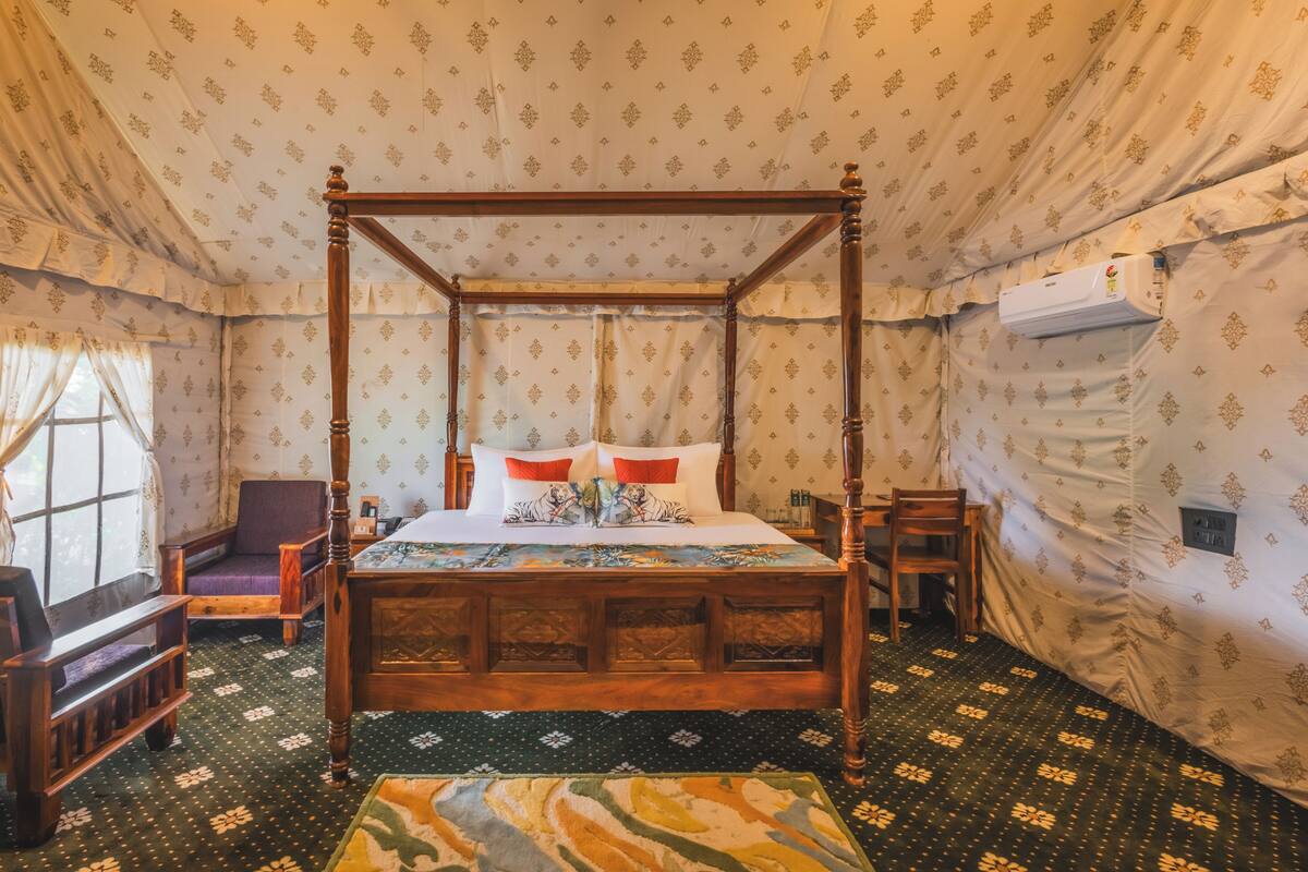A four-poster bed with a floral bedspread, a wooden bench, a chair, and a patterned carpet in a room with patterned walls and a window with curtains.