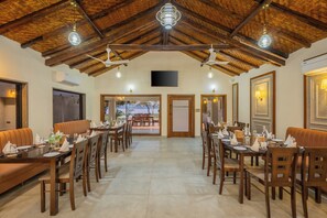 A dining area with wooden tables and chairs, a high ceiling with exposed beams, and a view through the door to an outdoor seating area. - The Forest Chapter By Sayaji Madhai (Sarangpur)