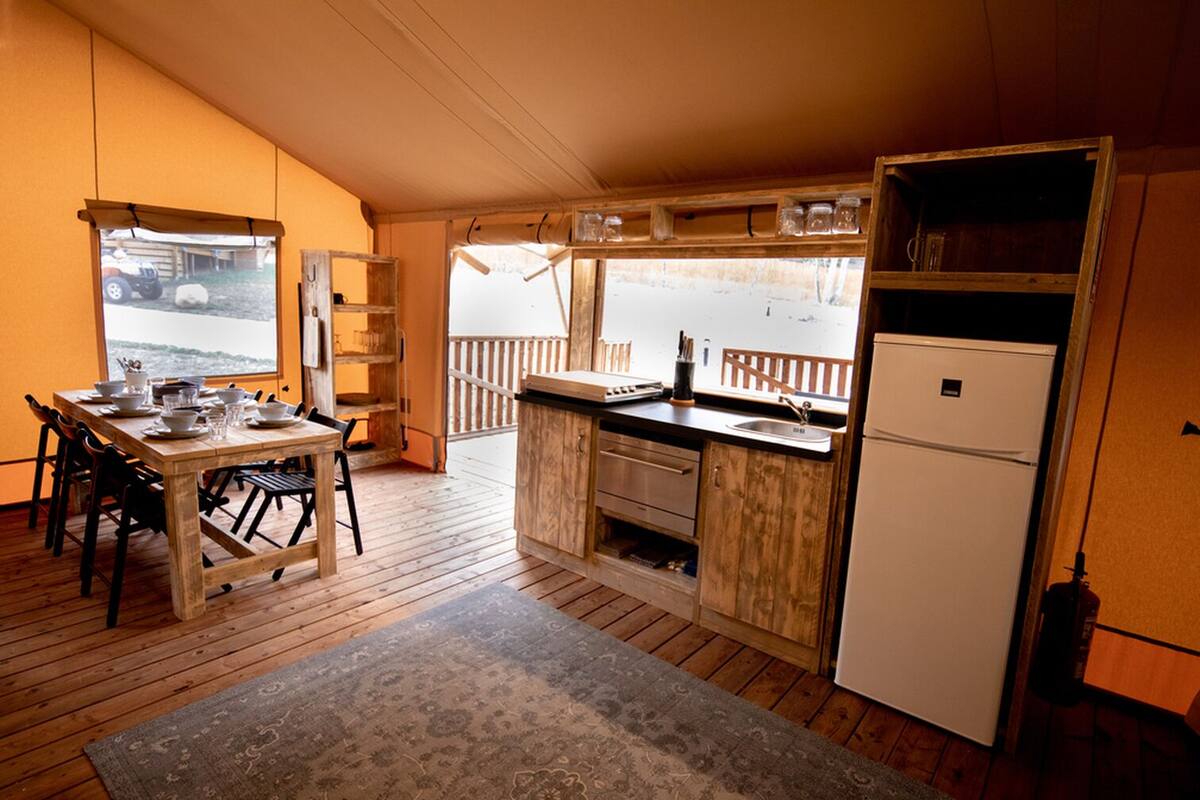 A compact kitchen with wooden cabinets, a dining table with chairs, a refrigerator, and a view of the outdoors through a window.