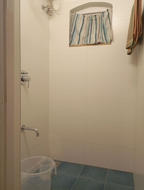 Un petit espace douche fermé, avec un mur recouvert de carreaux blancs, une pomme de douche, un porte-serviettes sur lequel est posée une serviette à rayures, et un sol recouvert de carreaux bleus.
