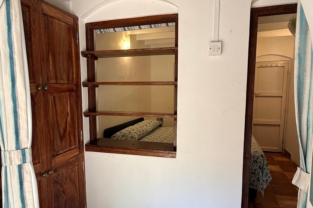 Una habitación con piso de baldosas, un armario de madera, una ventana con repisa y una puerta que da a otra habitación.