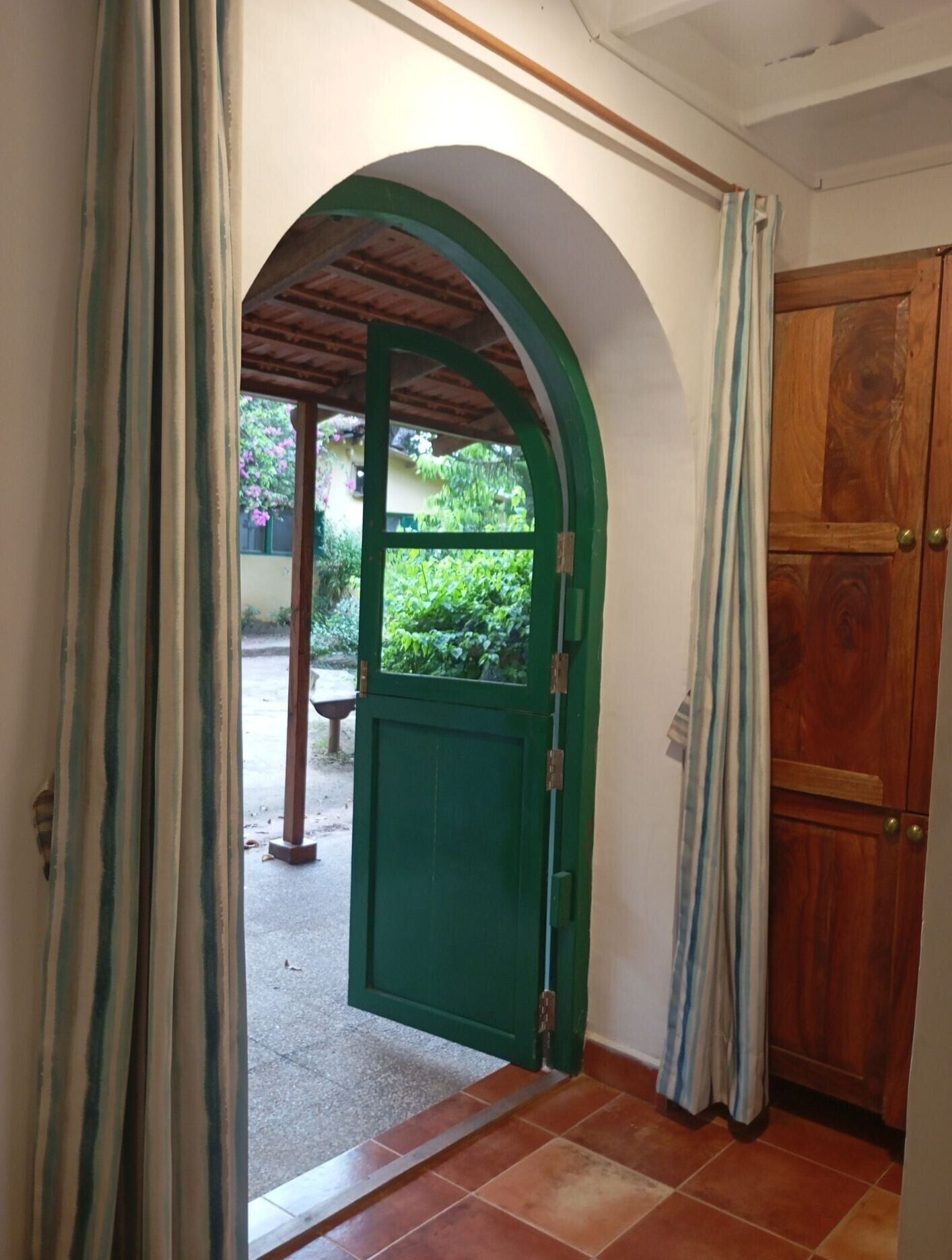Un arco verde y una puerta de madera conducen a un espacio al aire libre con un banco y follaje.