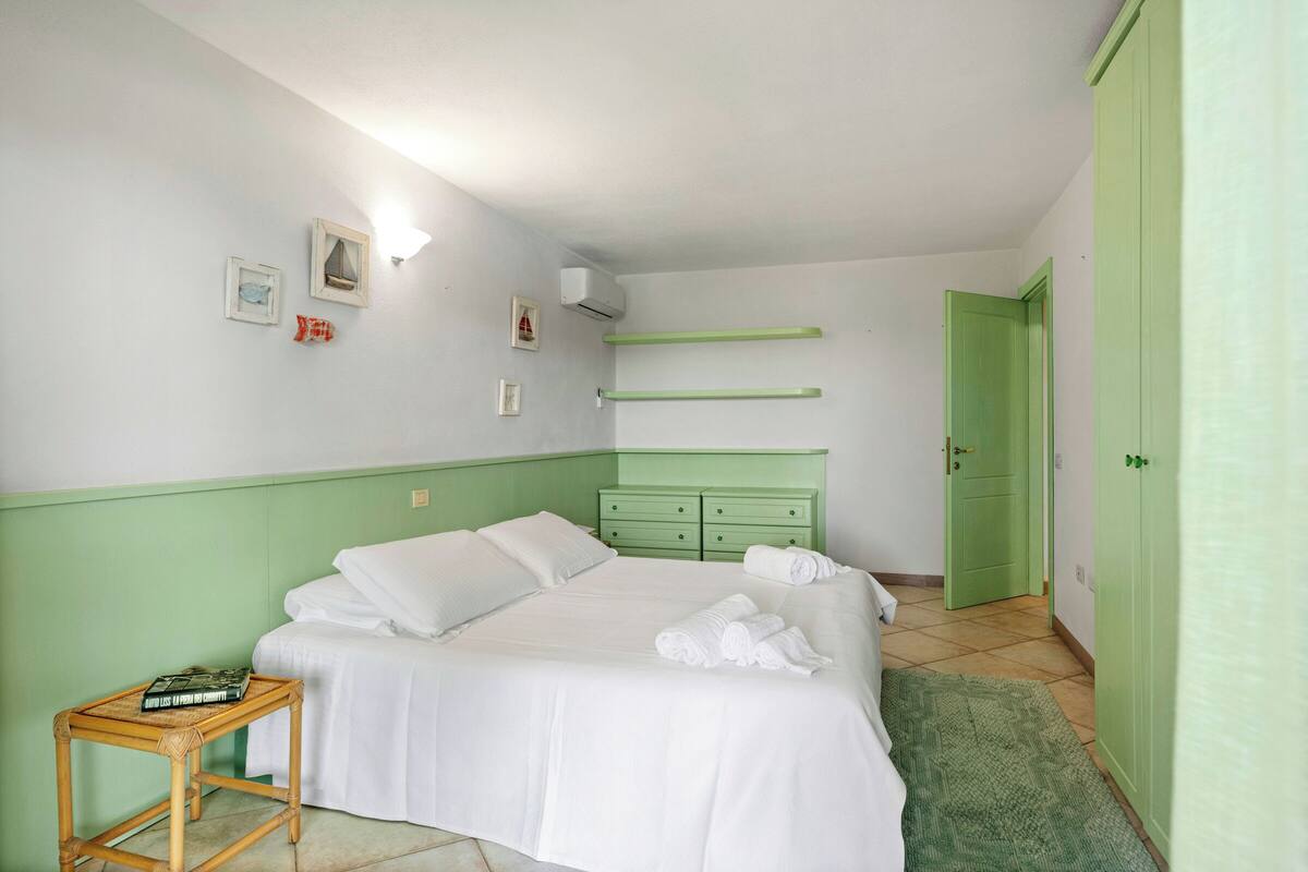 Een net opgemaakt bed met witte lakens, een nachtkastje met een boek, en een groene deur op de achtergrond.