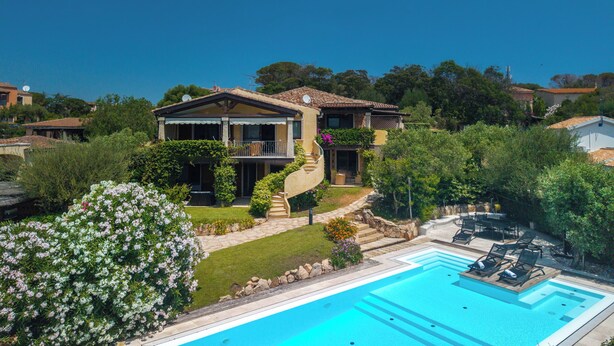 Una casa de dos pisos con piscina, rodeada de follaje y un cielo azul despejado.