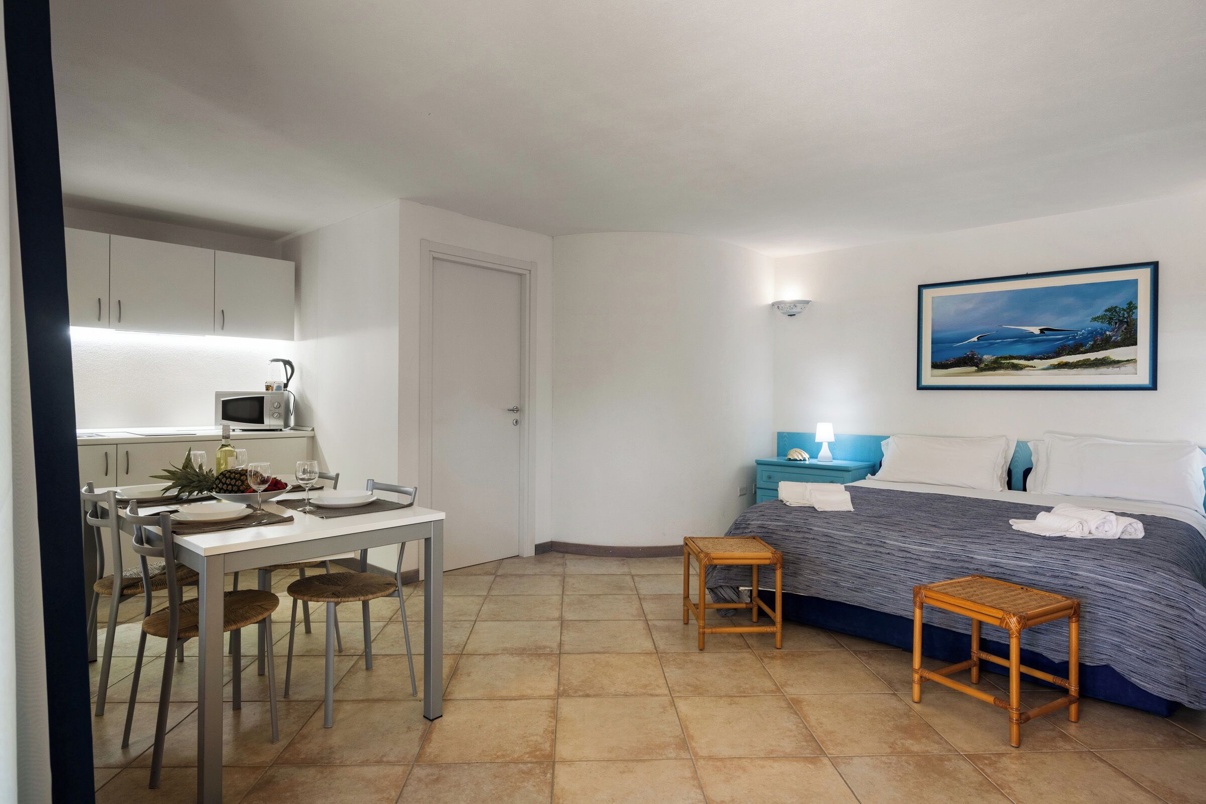 Habitació petita amb llit, taula, zona de menjador amb cadires, microones i un quadre a la paret.