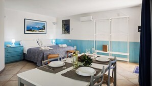Habitación de hotel con cama, una mesa de comedor para dos, pequeña cocina y ventana con cortinas.