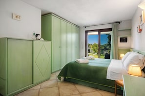 A bedroom with a bed, a green wardrobe, a window with a view, and a bedside table with a lamp.