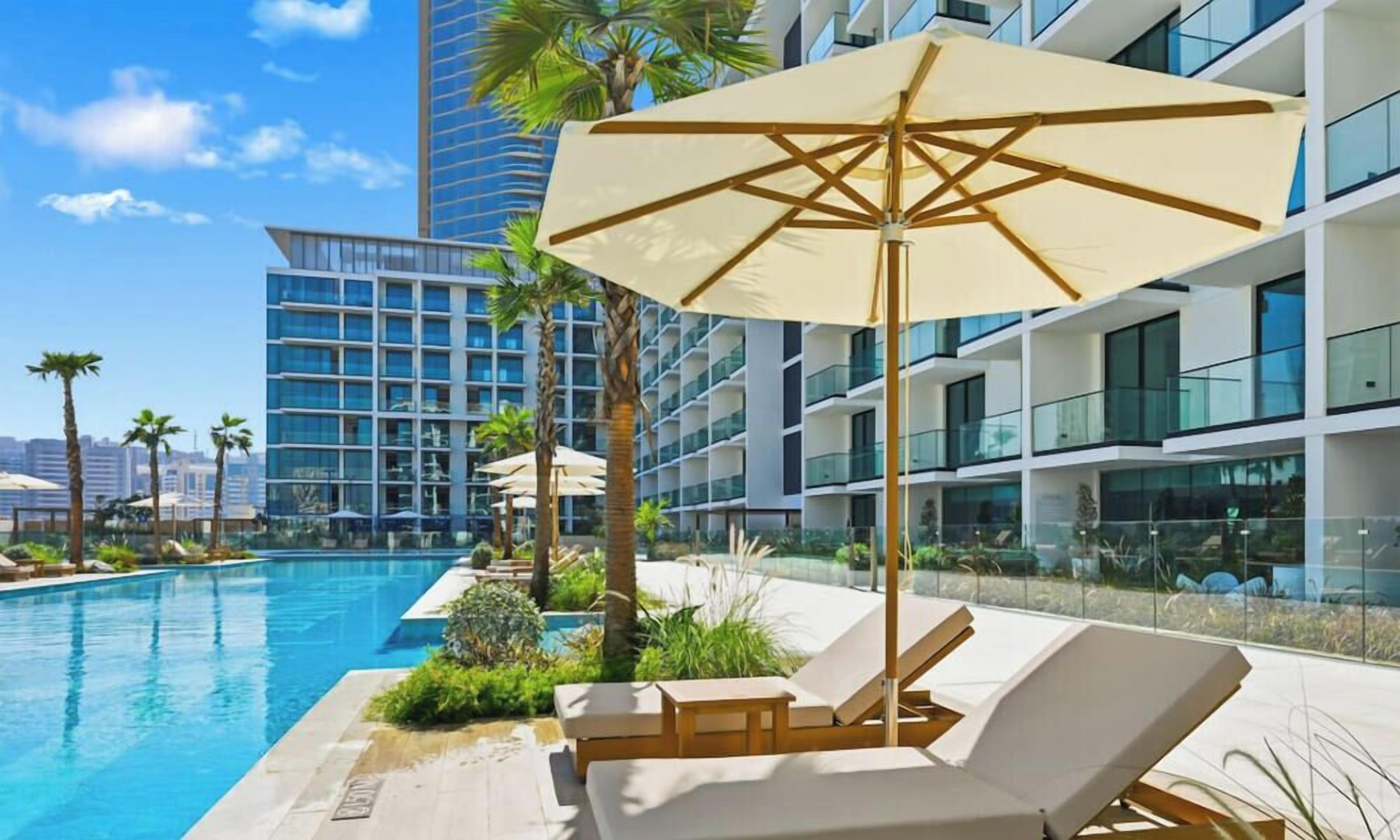 A modern hotel pool area with lounge chairs and umbrellas, surrounded by palm trees and a city skyline.