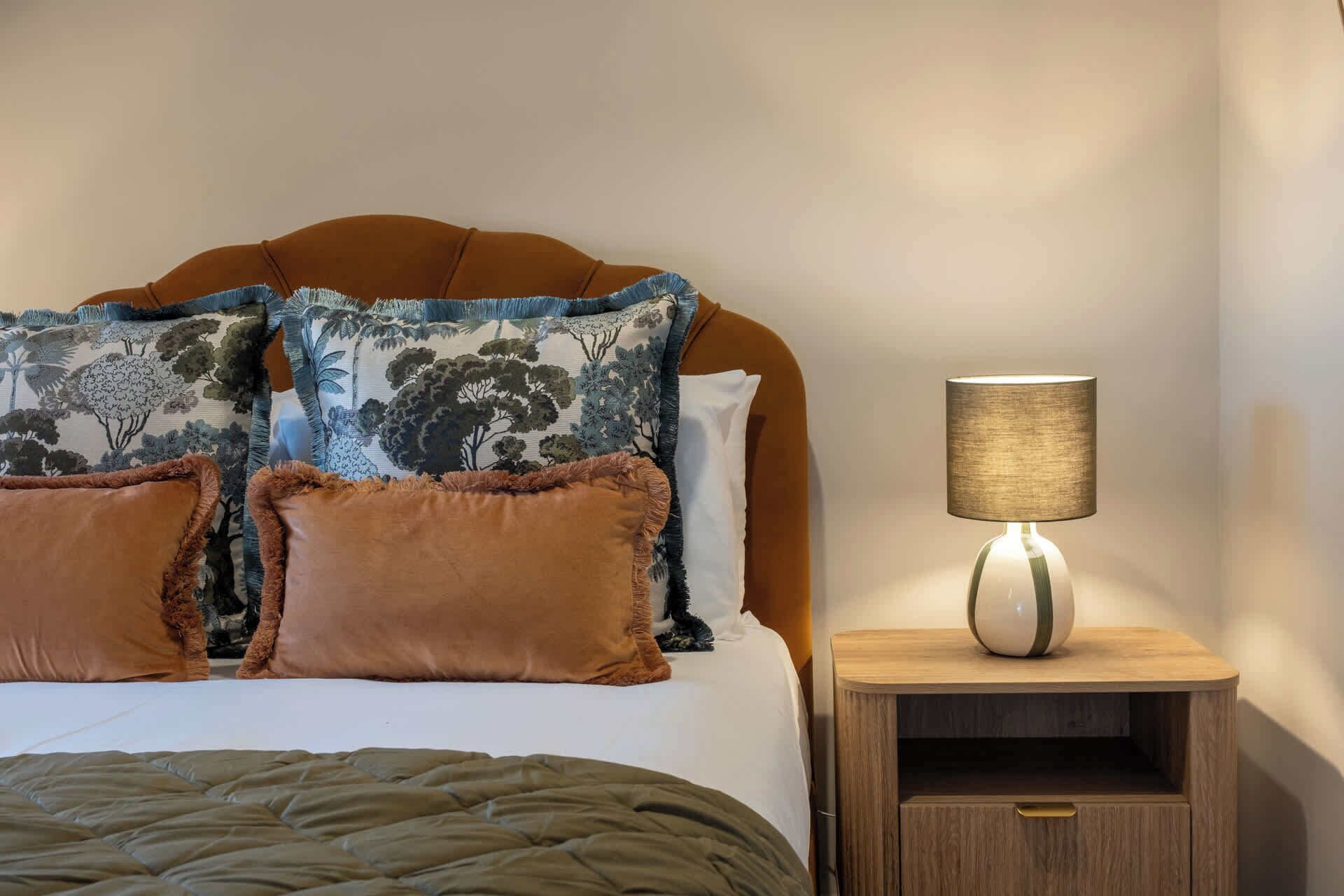 A neatly made bed with a brown headboard, two decorative pillows, a green blanket, and a bedside table with a lamp.