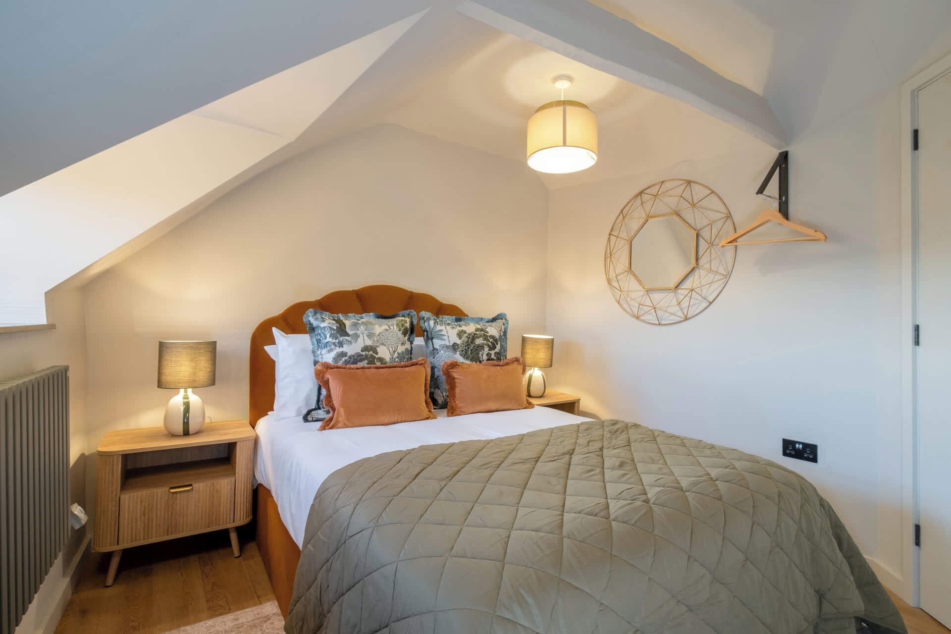Une chambre avec un plafond en pente, un lit recouvert d’une couette matelassée, deux lampes de chevet, une table de chevet en bois et un miroir fixé au mur.