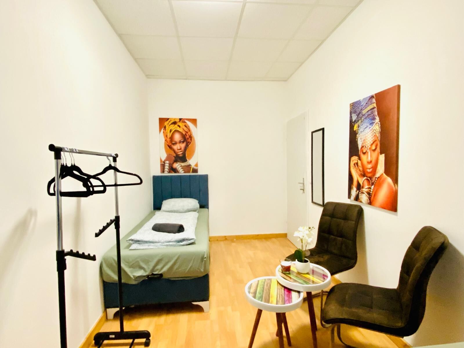 Ein kleines Hotelzimmer mit einem Einzelbett, einem Sessel, einem kleinen Tisch mit Pflanze und einem Kleiderständer.
