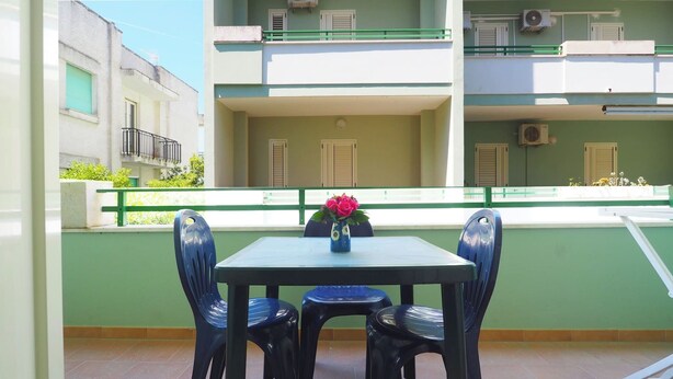 A balcony with a table and chairs, a vase with flowers, and a view of a building with shutters. - Robinia 2 Baia Verde - 3 Posti Letto - Posto Auto (Gallipoli)