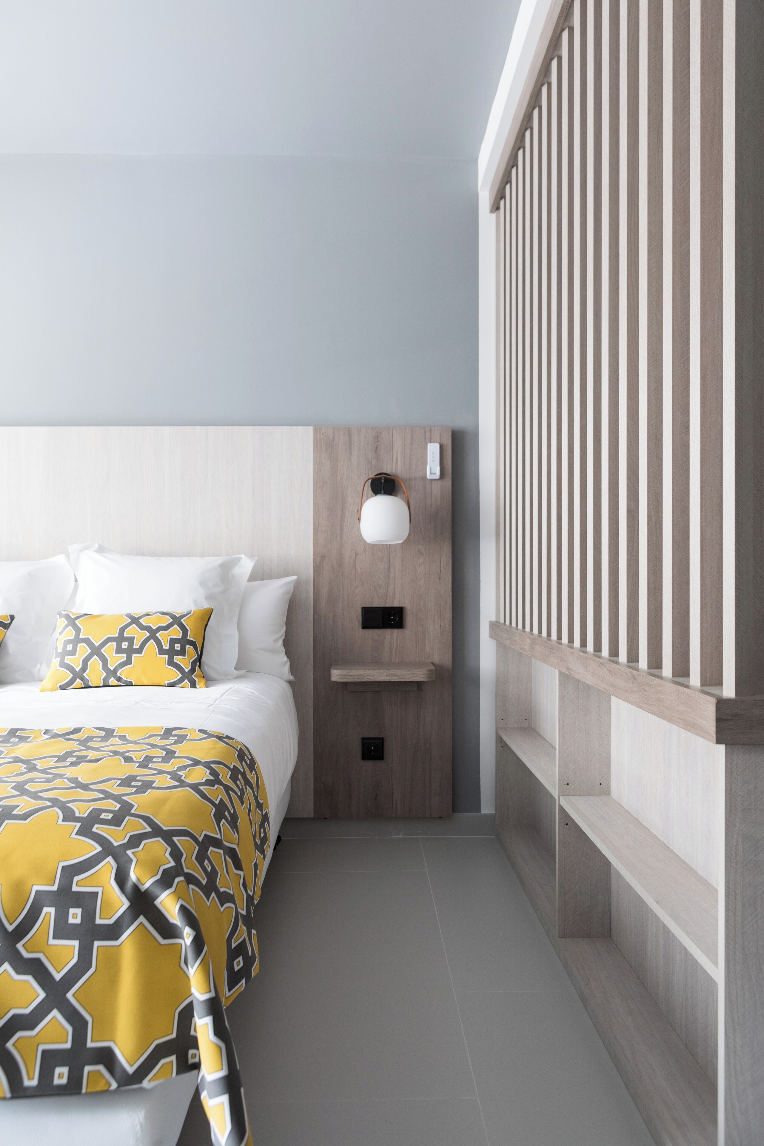 A modern hotel room with a bed, a nightstand, a wall-mounted lamp, and a wooden panel design.