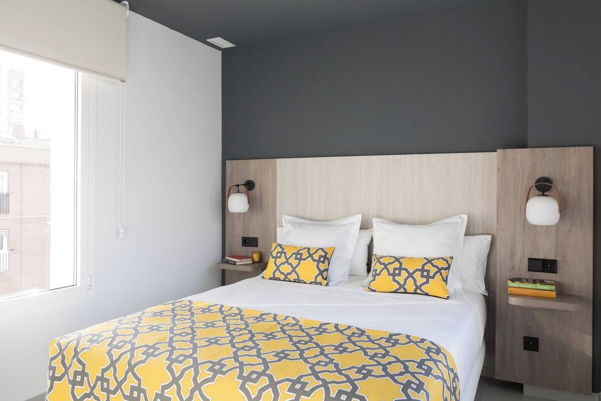 A modern hotel room with a large bed, two bedside tables with lamps, a window with blinds, and a view of buildings outside.