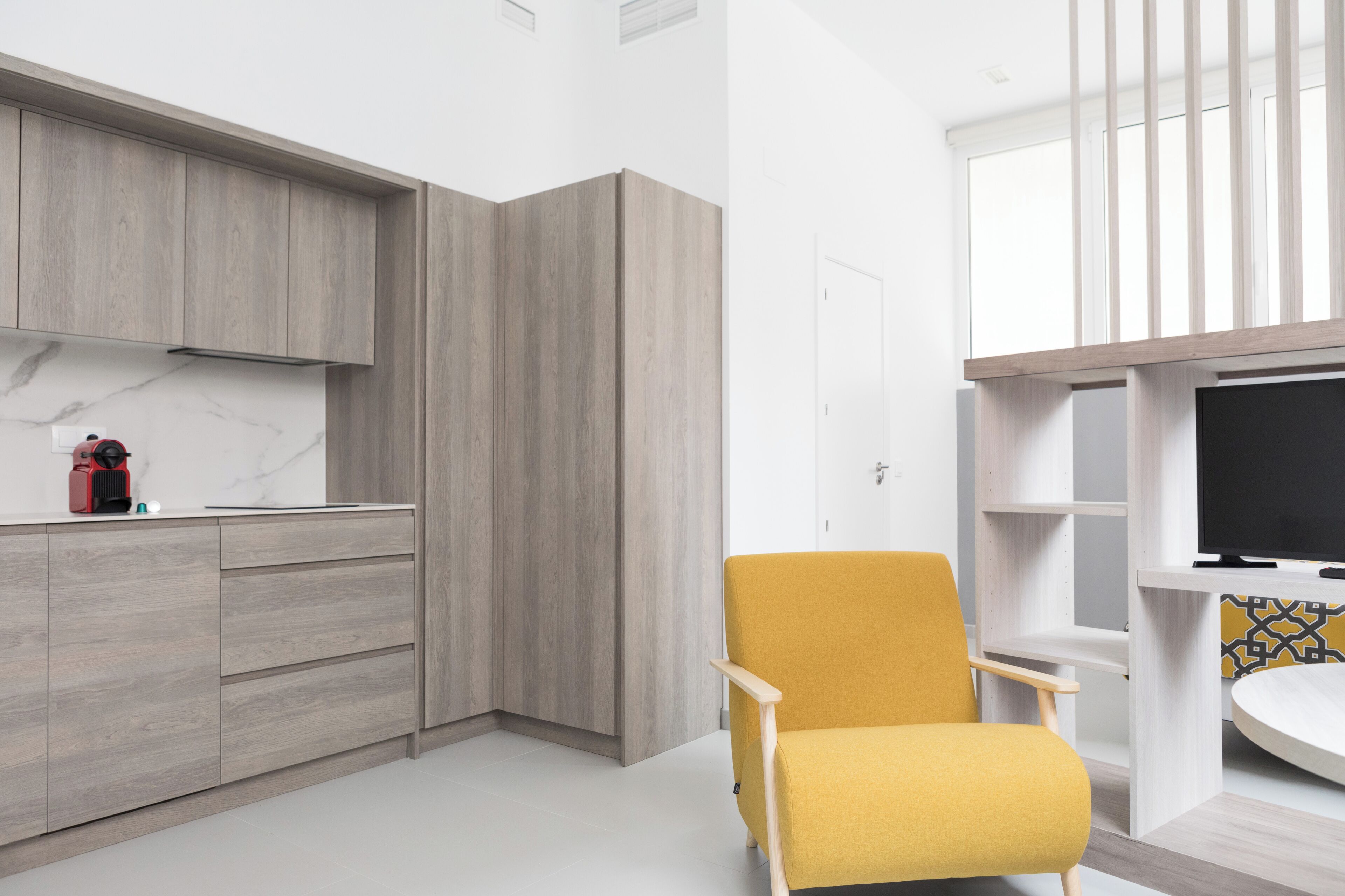 Una cucina moderna con mobili impiallacciati in legno, una poltrona gialla e una TV su uno scaffale bianco.