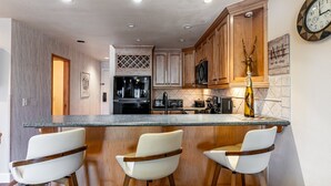 Cucina con mobili in legno, frigorifero in acciaio inossidabile, forno a microonde e angolo bar con tre sgabelli bianchi.