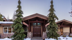 A two-story house with a stone foundation, a wooden front door, and two large evergreen trees flanking the entrance. - Summer by the Lake! Beautiful Swimming Pool, Patio Hot Tub Firetable BBQ Nature Trails Hiking Biking (Kamas)