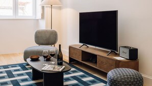 A living room with a large flat-screen TV, a striped armchair, a wooden coffee table with wine glasses and a bottle, a patterned rug, and a small ottoman. - Ando Living Abrantes 50 House - Deluxe Apartments (Lisbon)