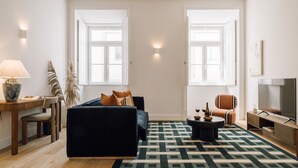 Un salon moderne avec un canapé foncé, une table basse et un fauteuil à rayures. On y trouve une lampe sur une table d’appoint, un téléviseur et une grande fenêtre qui laisse entrer la lumière naturelle.